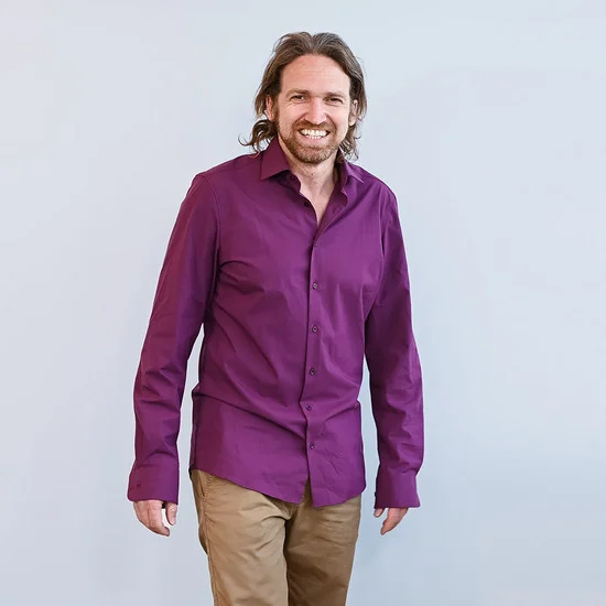 Ein Mann mit langen braunen Haaren und Bart lächelt, trägt ein lila Hemd und beige Hose.