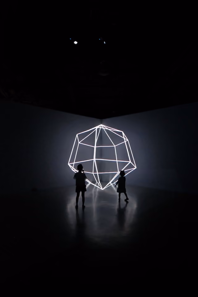 Zwei Personen stehen vor einem leuchtenden geometrischen Neon-Lichtobjekt in einem dunklen Raum.