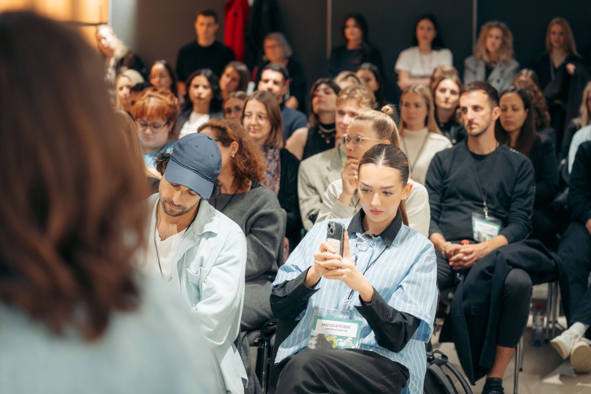 Menschen sitzen bei einer Konferenz; eine Frau im Vordergrund benutzt ihr Smartphone.