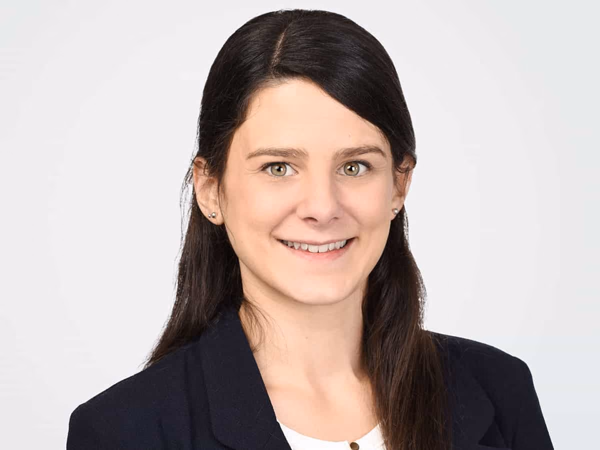 Porträt einer lächelnden Frau mit langen dunklen Haaren und dunklem Blazer vor hellem Hintergrund.