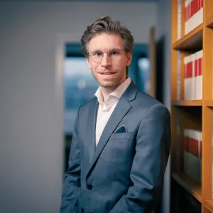 Junger Mann mit Brille, im Anzug stehend neben einem Bücherregal in einem Büroflur.