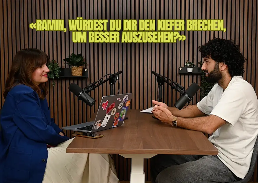 Zwei Personen sitzen an einem Holztisch mit Mikrofonen für ein Gespräch oder Podcast.