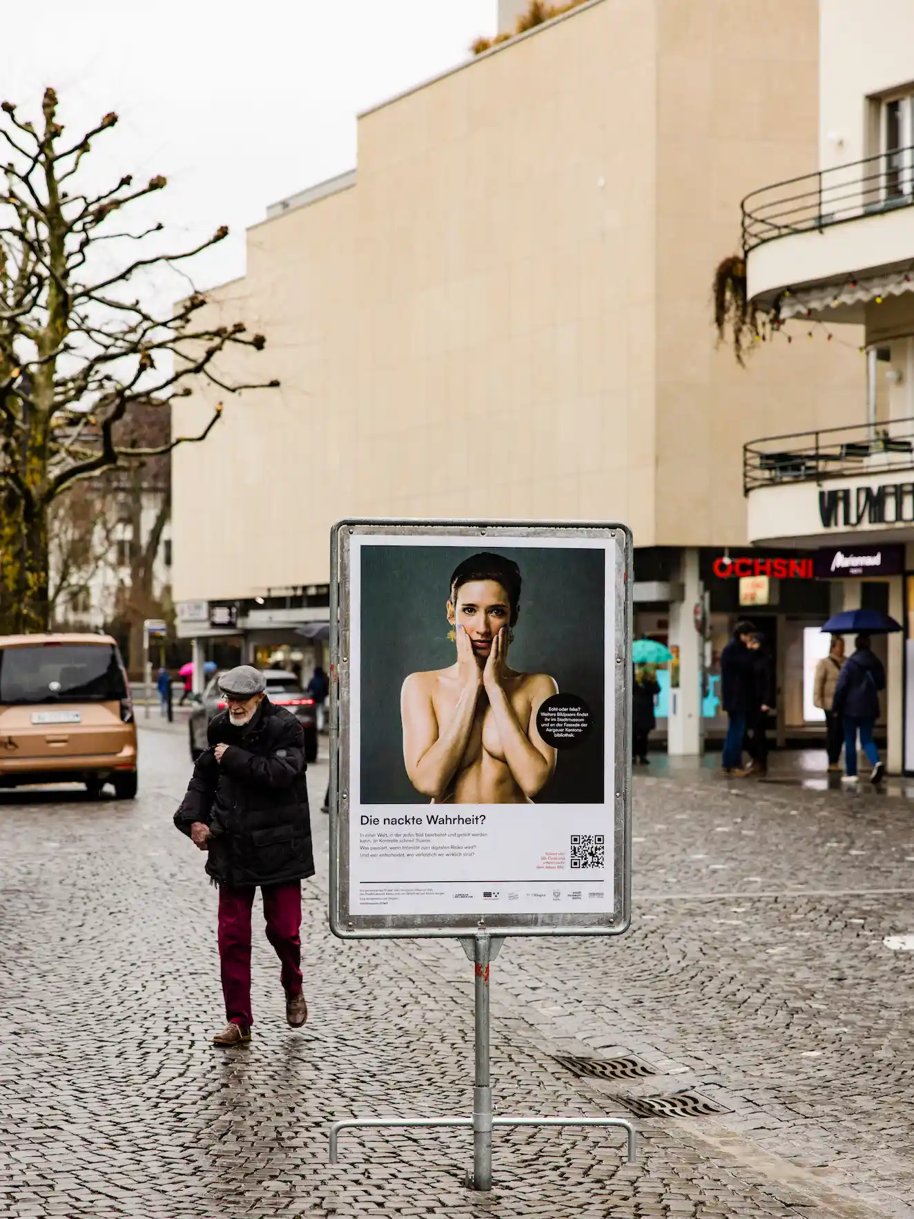 Plakat mit nackter Frau und dem Titel „Die nackte Wahrheit?“ auf Kopfsteinpflaster in Stadt.