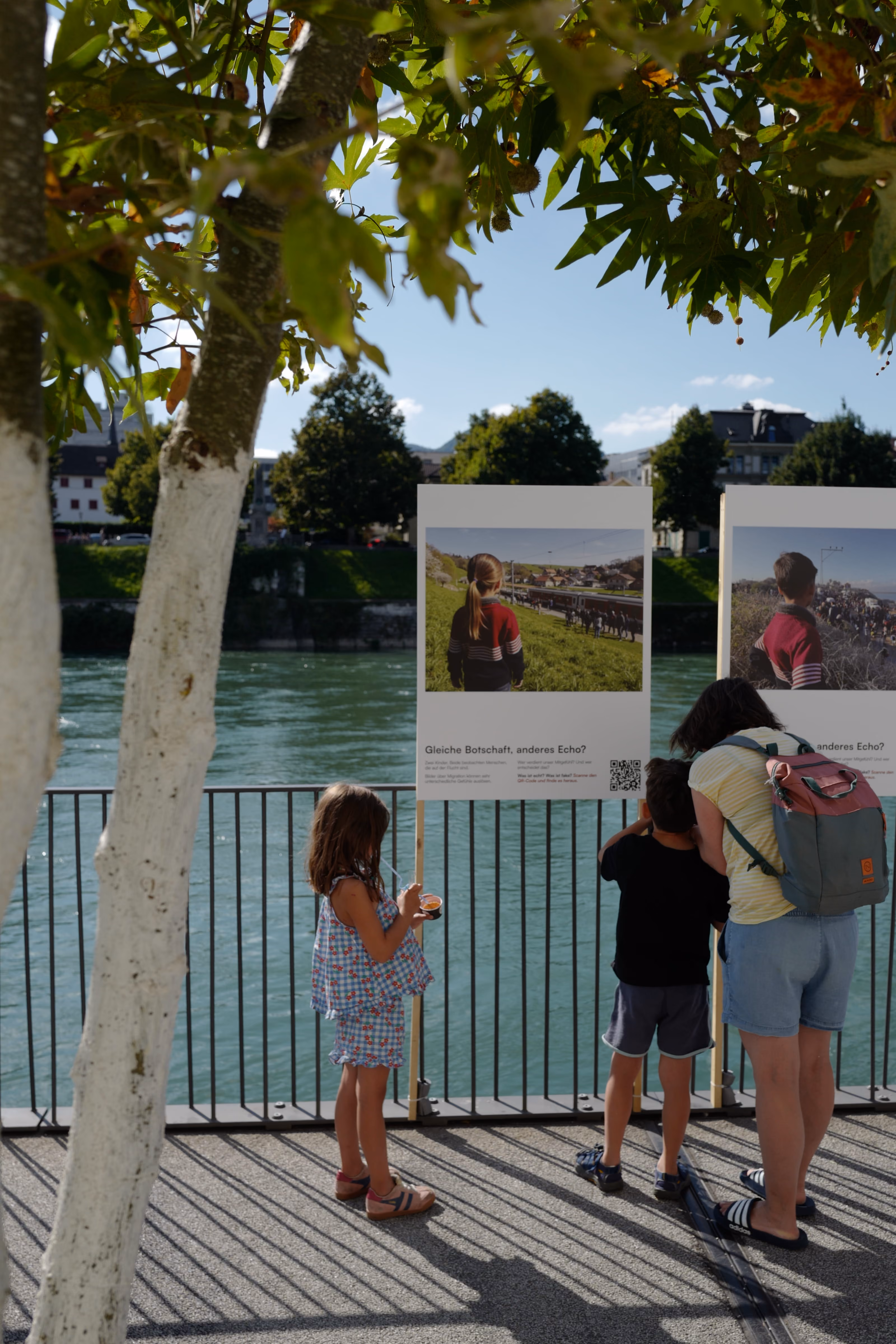 Frau mit zwei Kindern liest draußen an einem Fluss vor Schildern mit Fotos und Text.
