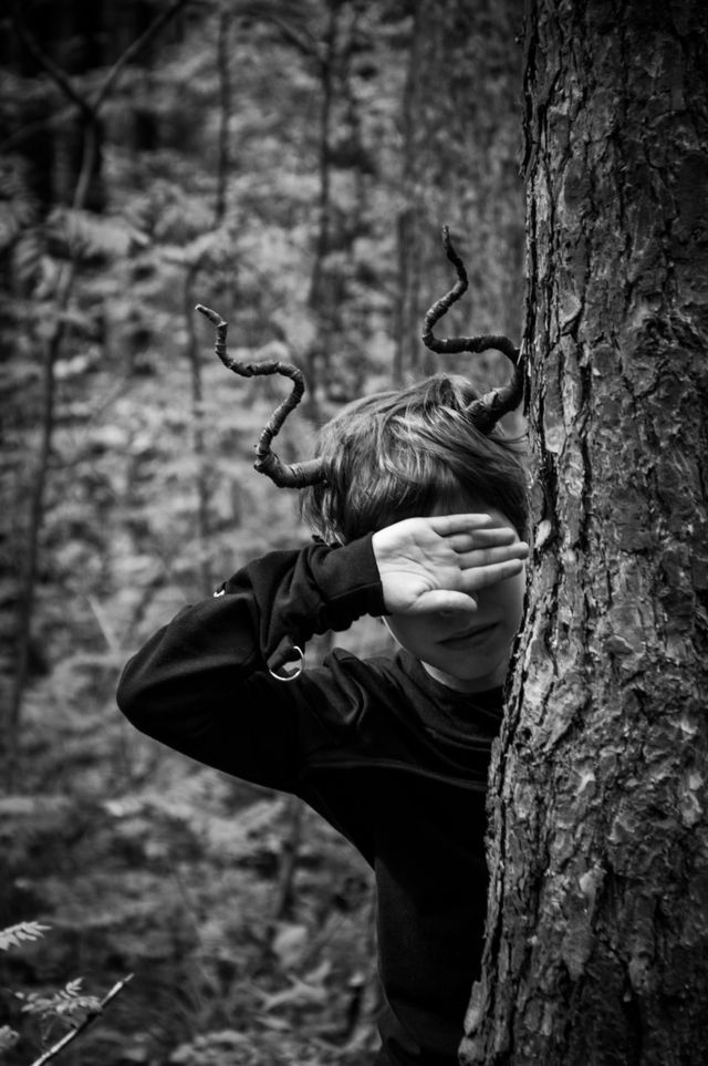 Kind mit verdrehten Ästen wie Hörner hinter einem Baum, Gesicht von Hand teilweise verdeckt.