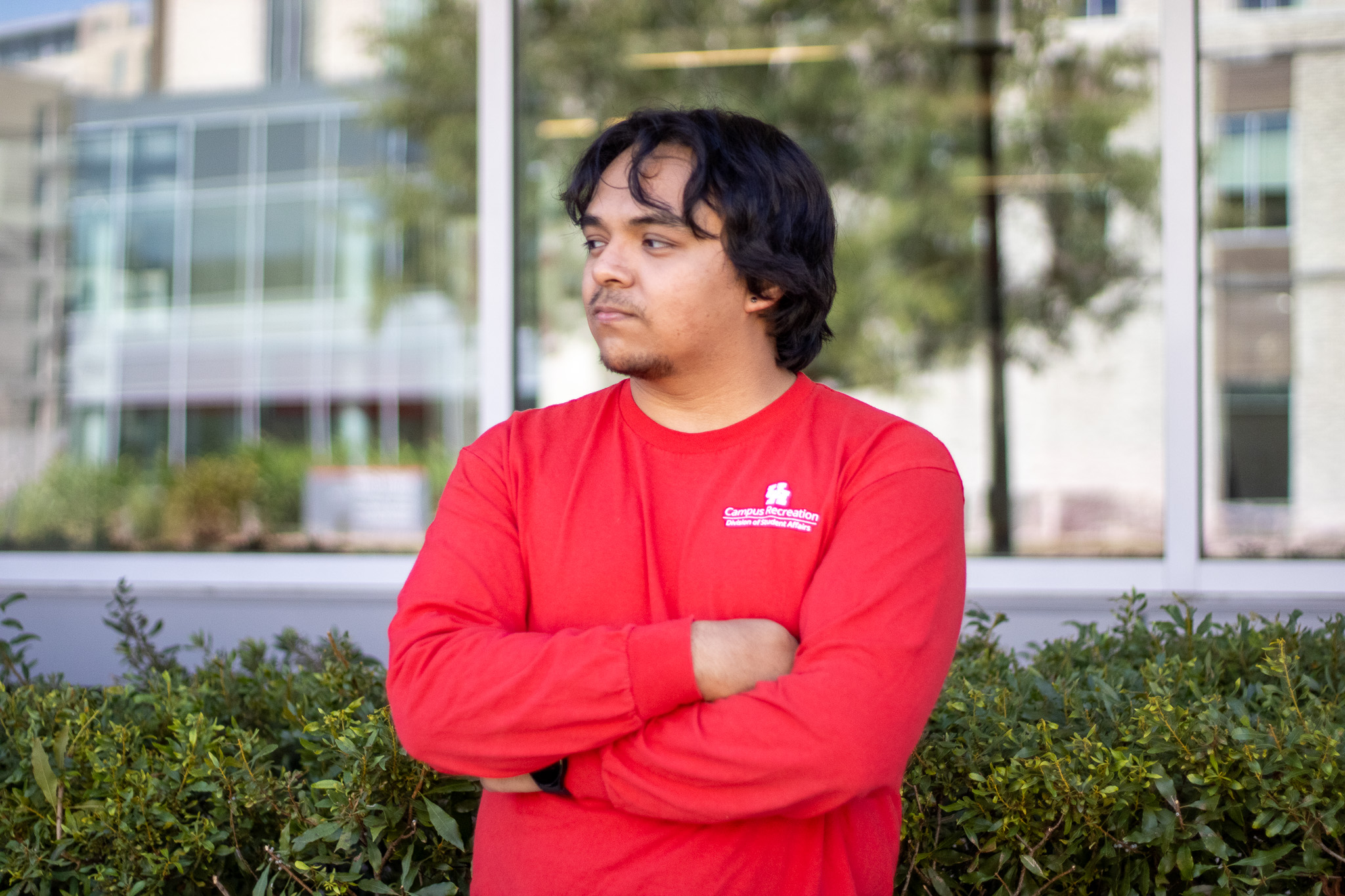 Young man in a red long-sleeve shirt with arms crossed standing in front of a building with windows and greenery.