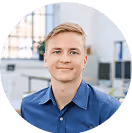 Junger Mann mit blauer Hemd lächelt in einem modernen Büro vor einem verschwommenen Hintergrund.