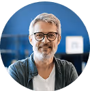 Älterer Mann mit Brille und grauem Haar lächelt freundlich, sitzt in modernem Büro, blauer Hintergrund.