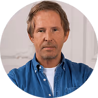 Mann mit Jeanshemd schaut ernst in die Kamera, im Hintergrund weiße Wand mit dekorativem Detail.