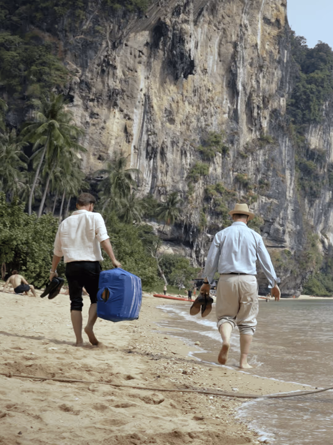 Zwei Männer spazieren am Strand, tragen Schuhe und Koffer, beeindruckende Felskulisse im Hintergrund.