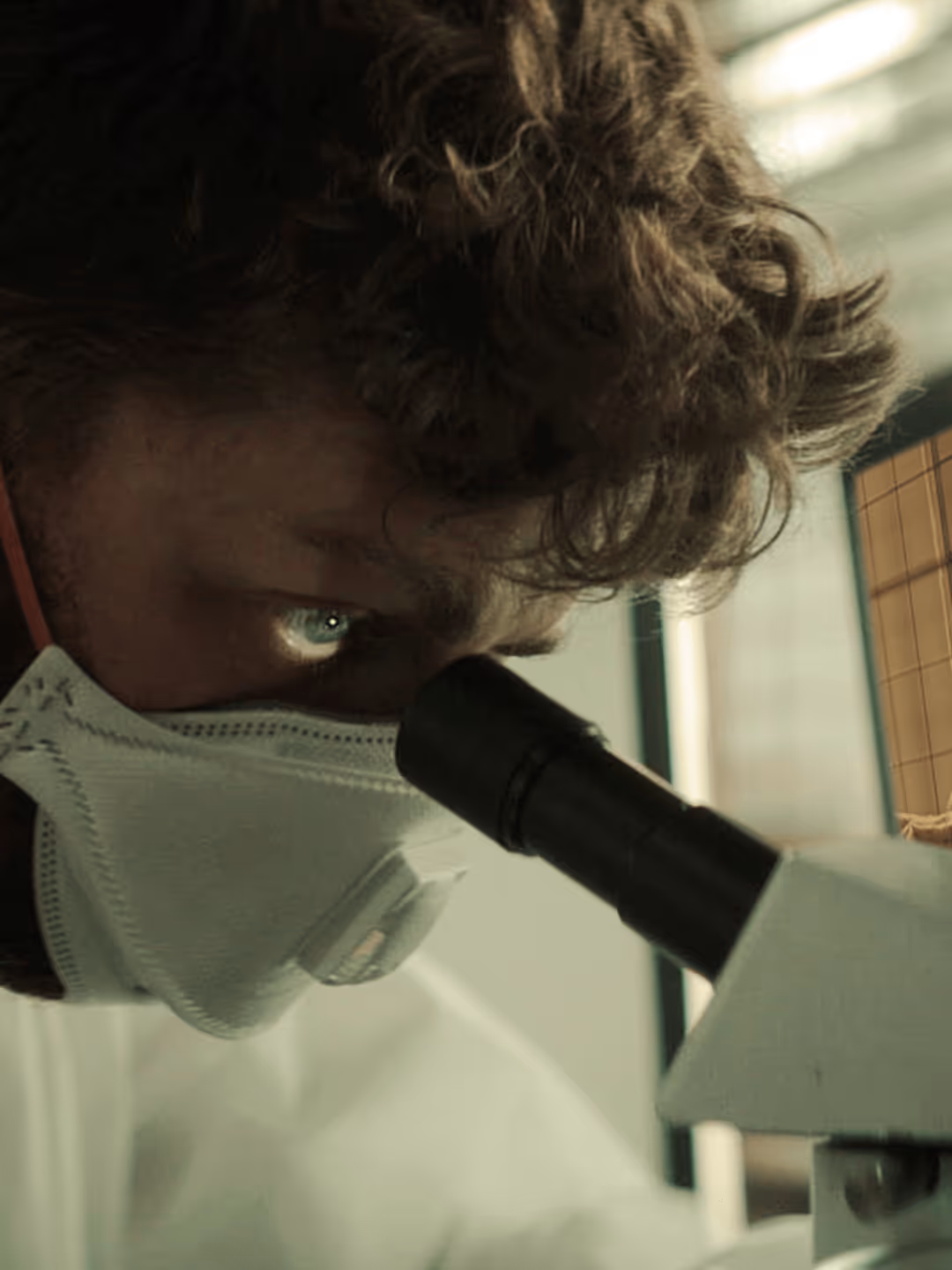 Wissenschaftler mit Schutzmaske blickt durch Mikroskop im Labor.