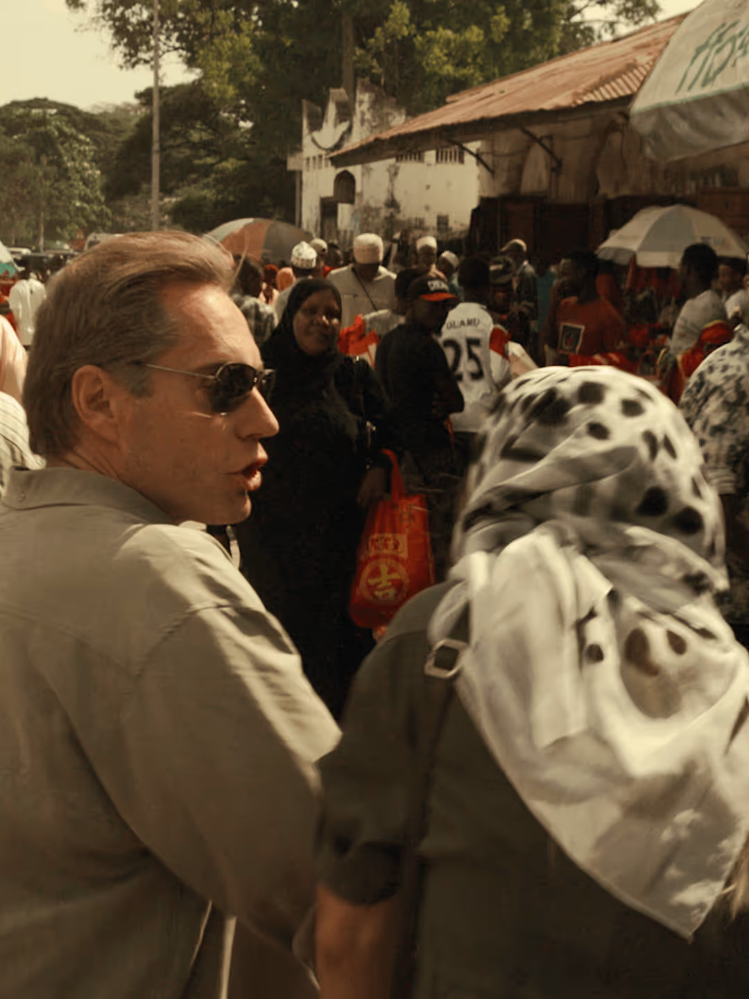 Belebter Markt mit diversen Menschen, einige tragen traditionelle Kleidung; ein Mann im Vordergrund, Gespräch mit verschleierter Frau.