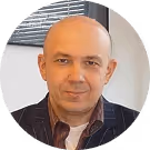 Glatzköpfiger Mann in gestreiftem Sakko lächelt leicht, vor einem Fenster in modernem Büro.