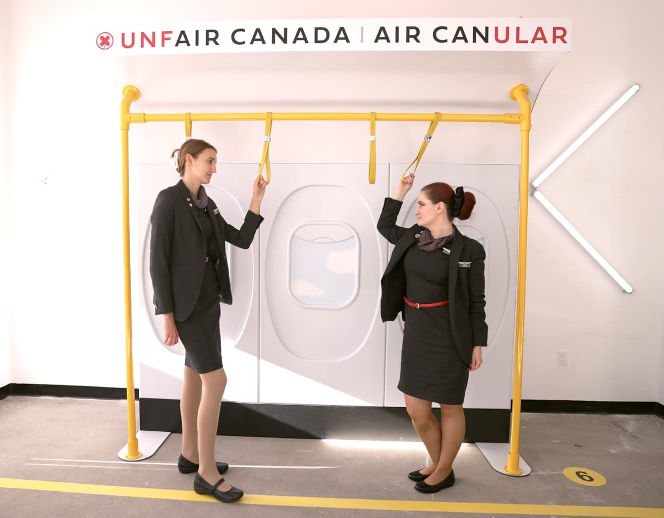 Two women dressed as flight attendants stand in a mock airplane interior with bus-style standing straps.