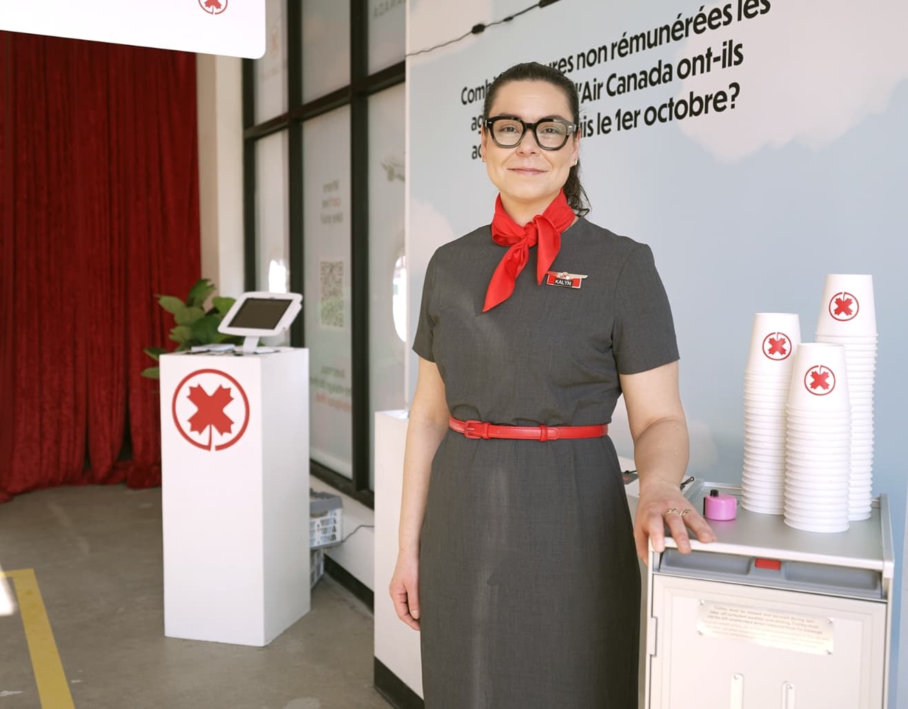 Woman dressed as a flight attendant stands beside a service cart with stacks of small paper cups.