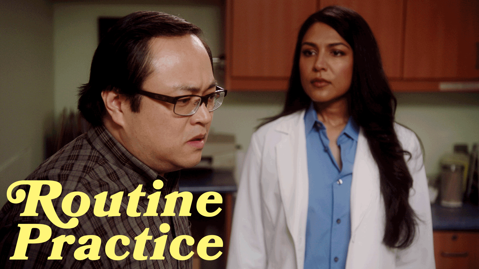 Man wearing glasses and a plaid shirt looks concerned while a woman in a white coat and blue shirt stands behind him in a clinical setting.