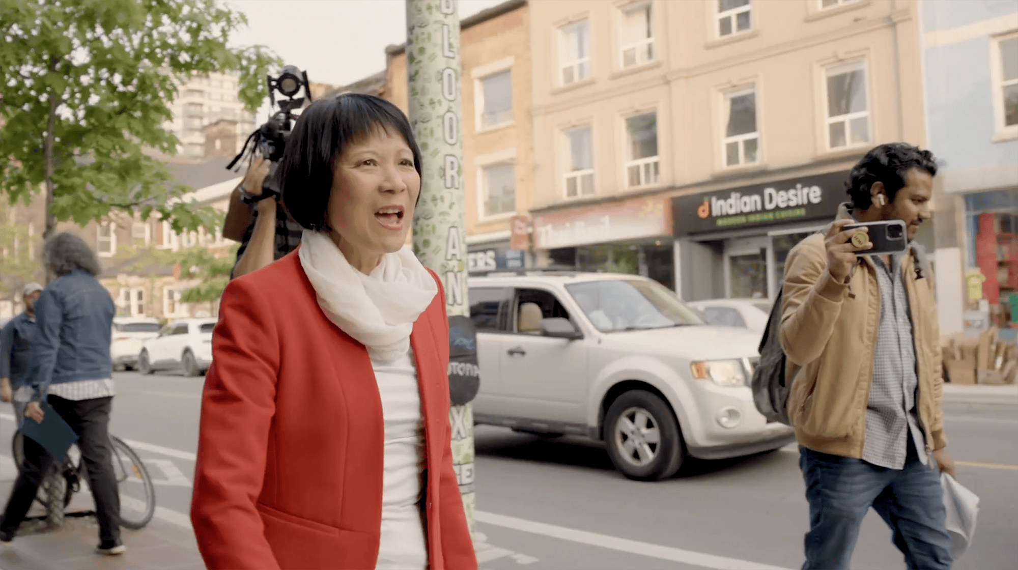 Woman in red jacket and white scarf speaking on a city street with a man holding a smartphone nearby and a white SUV and store signs in the background.