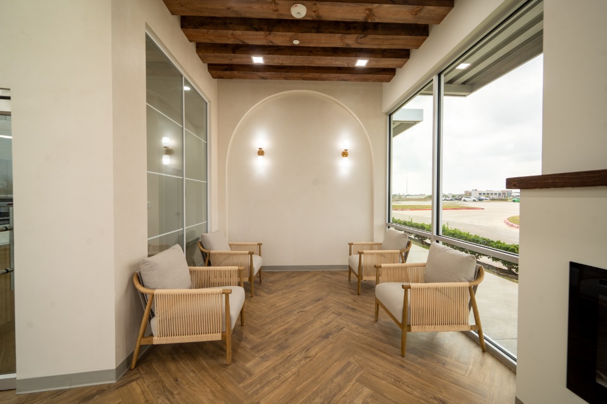 Modern waiting area with four beige cushioned wooden chairs, wood beam ceiling, and large windows overlooking a parking lot.