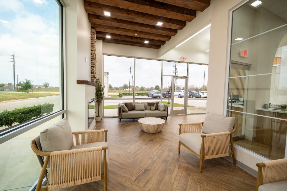 Modern lobby area with large windows, wooden ceiling beams, light wood flooring, a grey sofa, two armchairs, and a round wooden coffee table.