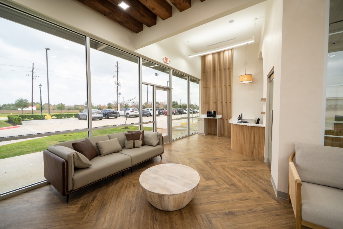 Modern lobby with large floor-to-ceiling windows, a beige and brown sofa with pillows, a round wooden coffee table, wood paneled floor and ceiling beams, a reception desk, and a coffee station.