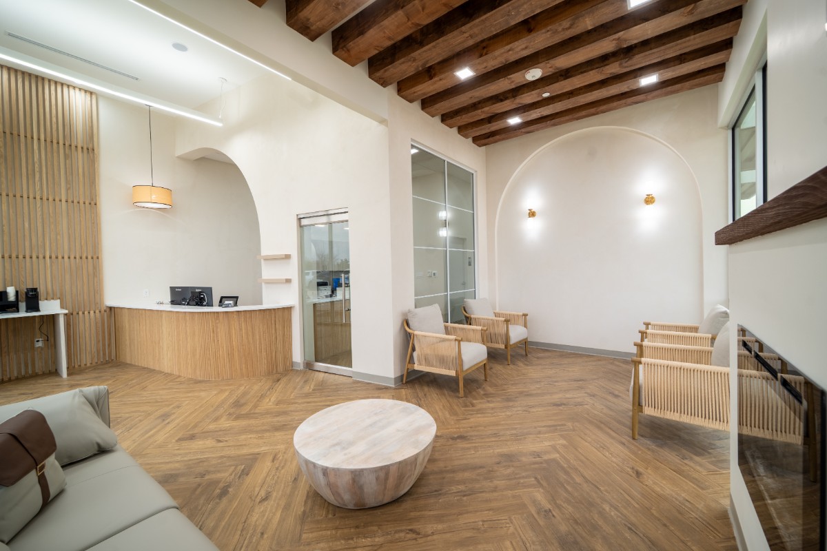 Modern waiting area with wooden floor, beige walls, cushioned chairs, a curved reception desk, and exposed wooden ceiling beams.