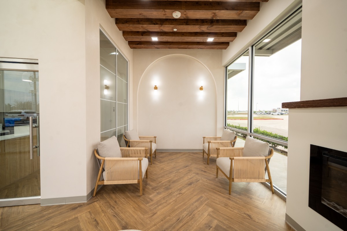 Modern waiting area with four wooden armchairs with beige cushions, wooden ceiling beams, and large windows overlooking a parking lot.