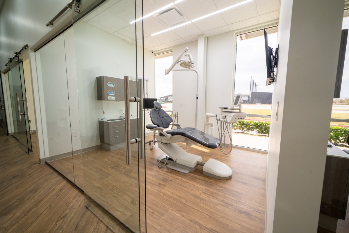 Modern dental treatment room with a reclining dental chair, overhead light, medical equipment, wood flooring, and large windows with a view outside.