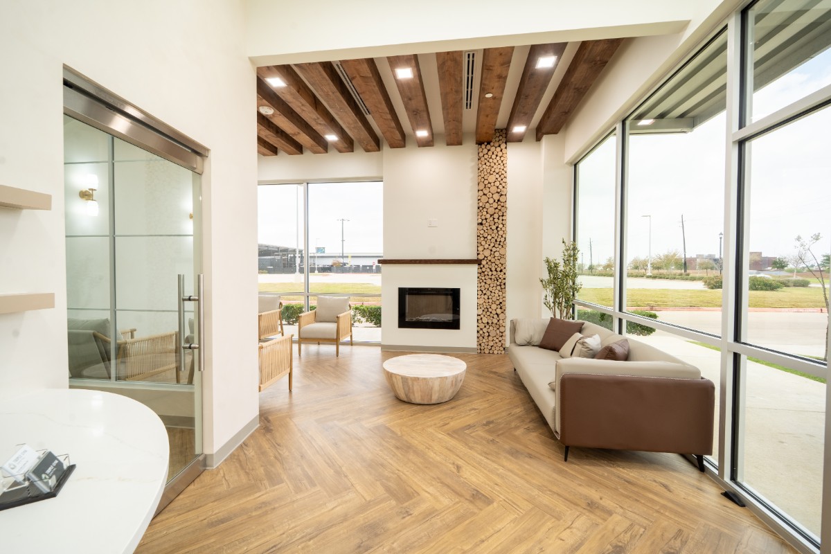 Bright modern living area with wood floor, wood beam ceiling, large windows, a fireplace, a beige sofa with brown accents, two armchairs, and a round wooden coffee table.