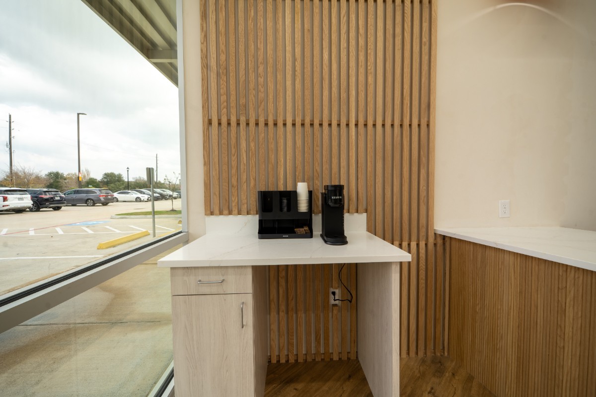 Minimalist coffee station with a Keurig coffee maker, disposable cups, and a wooden slatted wall, next to a large window showing parked cars outside.