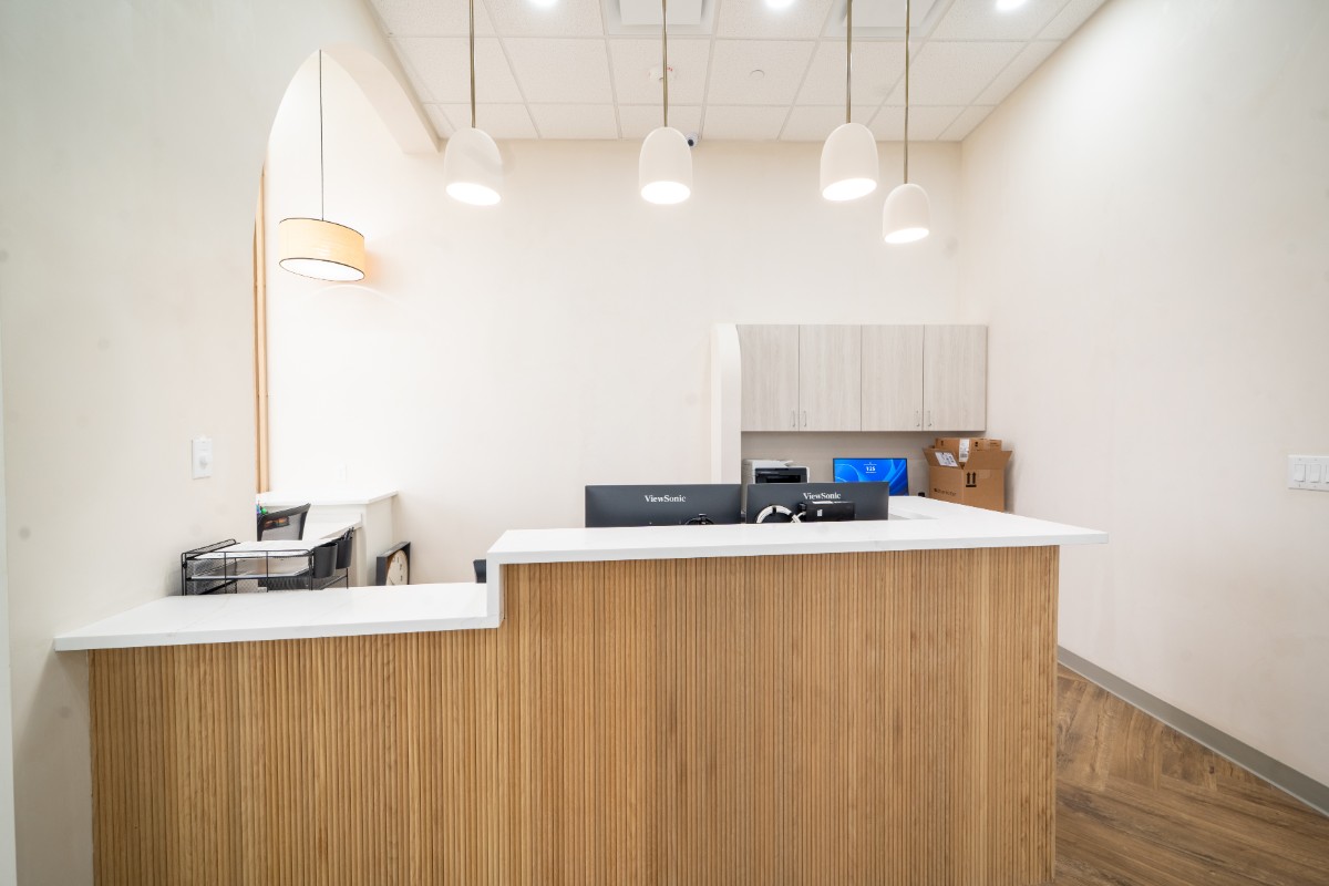 Modern reception desk with white countertop, wooden base, three pendant lights, and computer monitors in a bright office.
