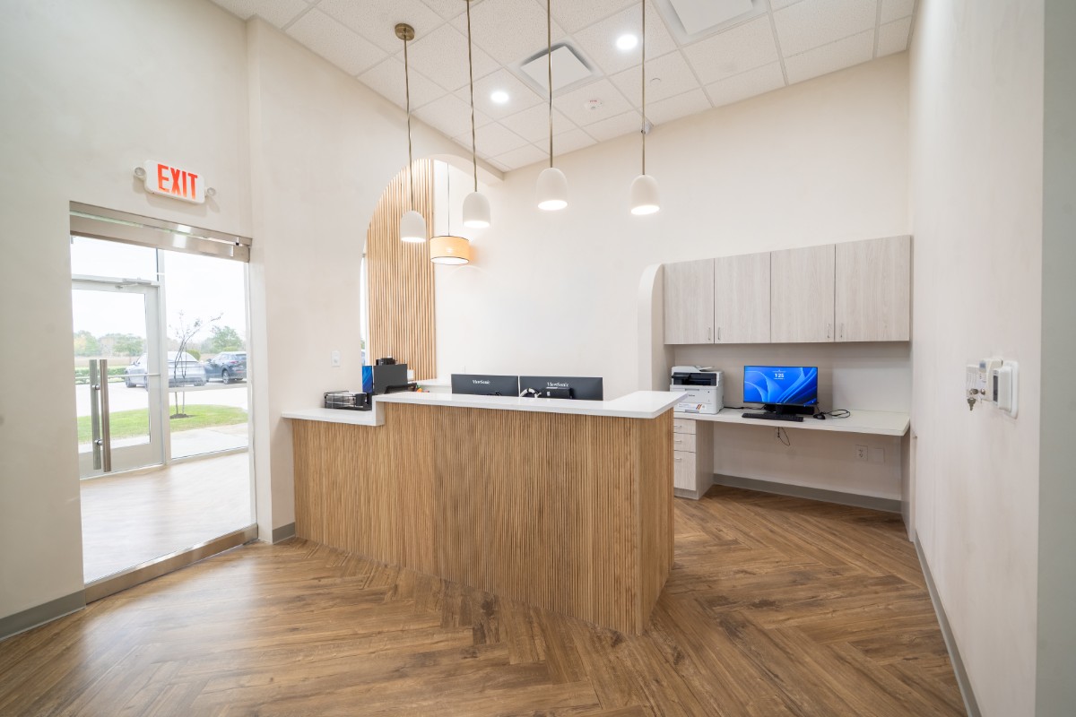 Modern office reception area with wooden desk, hanging pendant lights, computer monitors, and a glass exit door.