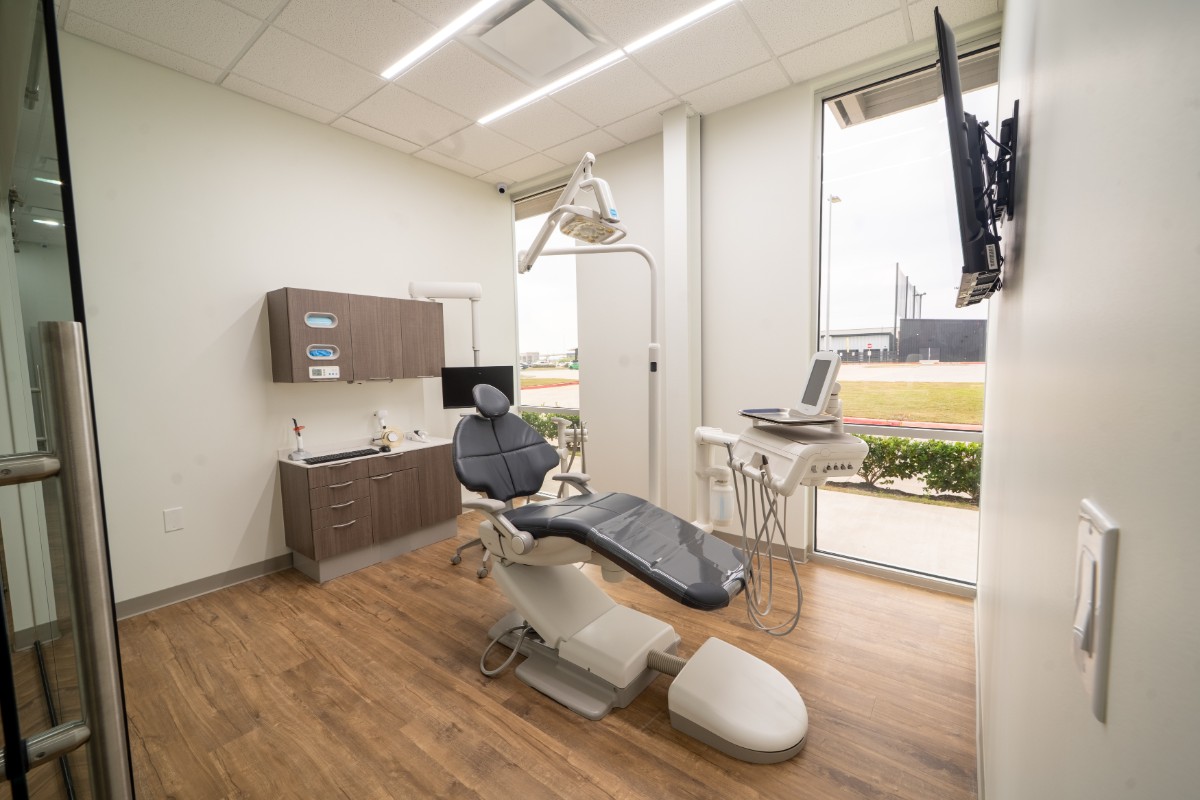 Modern dental clinic room with a dental chair, overhead lamp, cabinets, computer monitor, and large window overlooking an outdoor area.