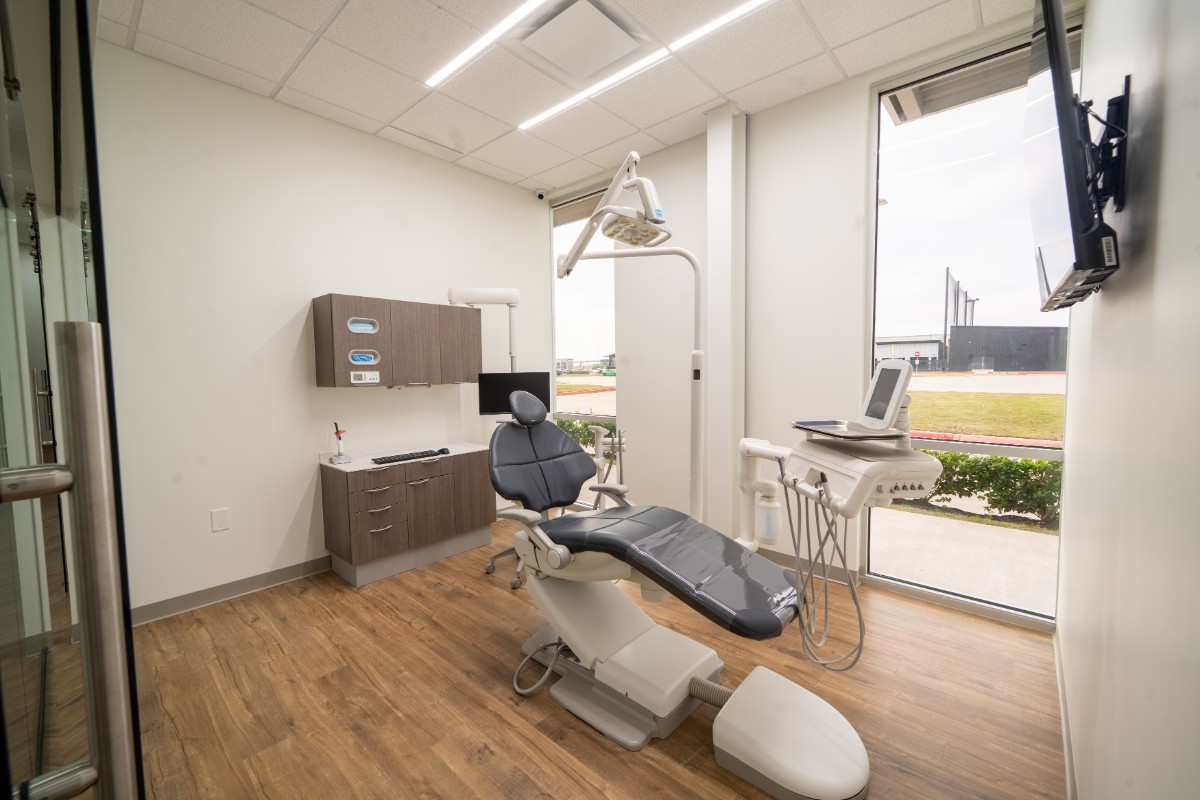 Modern dental examination room with black dental chair, overhead light, equipment, cabinets, and large windows overlooking outdoor area.
