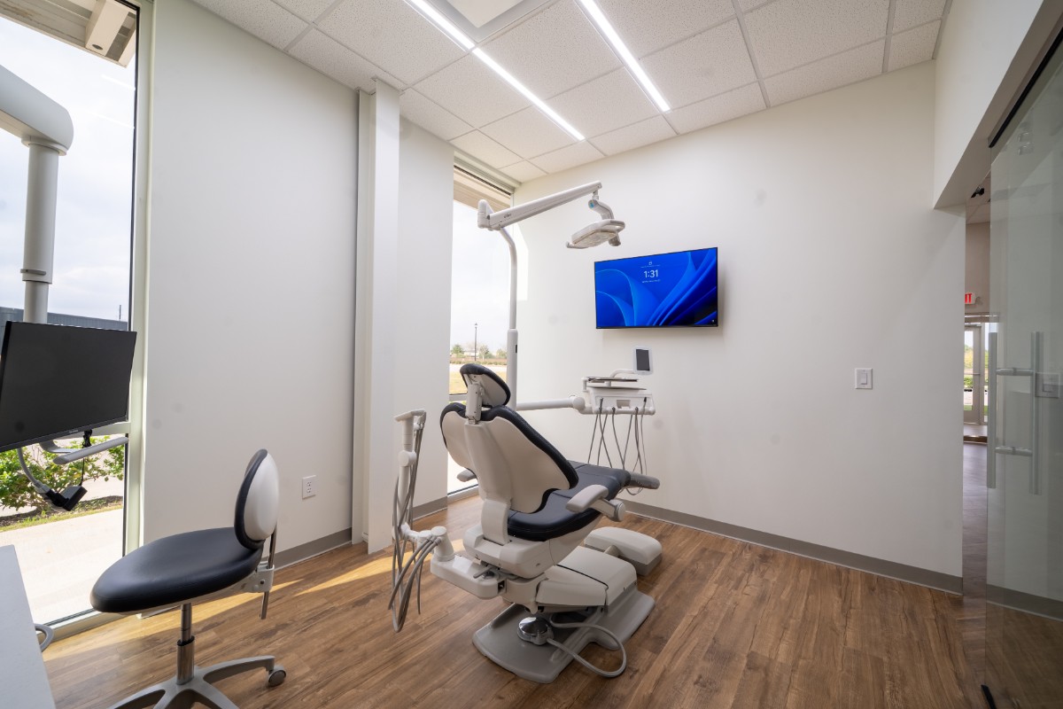 Modern dental exam room with dental chair, equipment, a wall-mounted monitor, and wooden flooring.