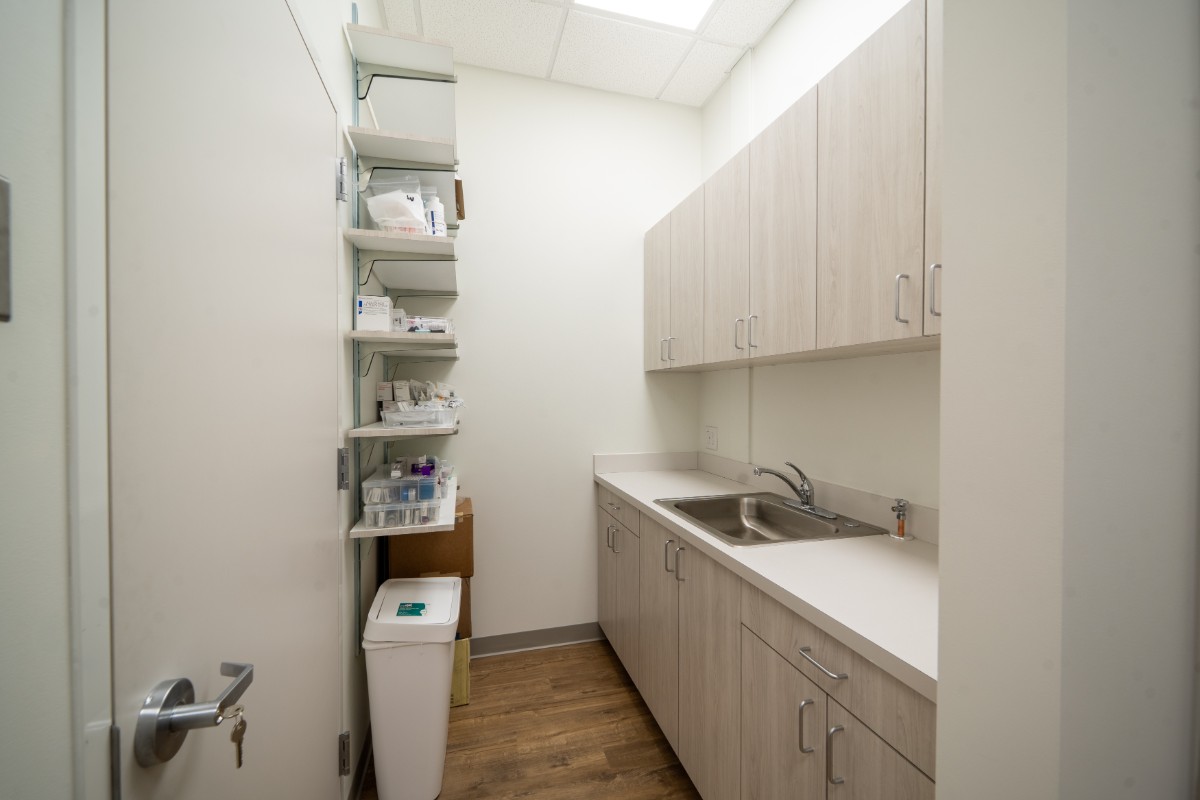 Small clean utility room with light wood cabinets, a stainless steel sink, white countertop, shelving with medical supplies, and a white trash bin.