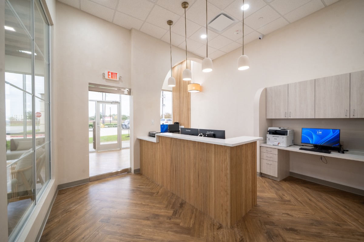 Bright modern office reception area with wooden desk, hanging pendant lights, and a workstation with a computer and printer.
