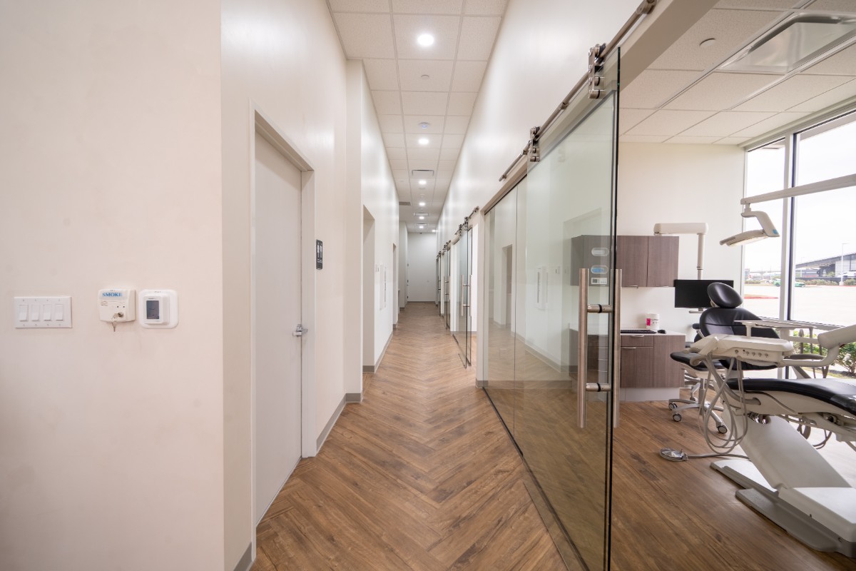 Modern dental clinic hallway with wooden floor, white walls, and glass doors leading to treatment rooms with dental chairs.