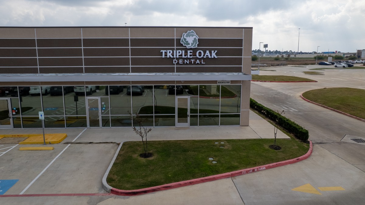 Front view of Triple Oak Dental clinic with reflective glass doors and windows, surrounded by parking spaces and a small landscaped area.