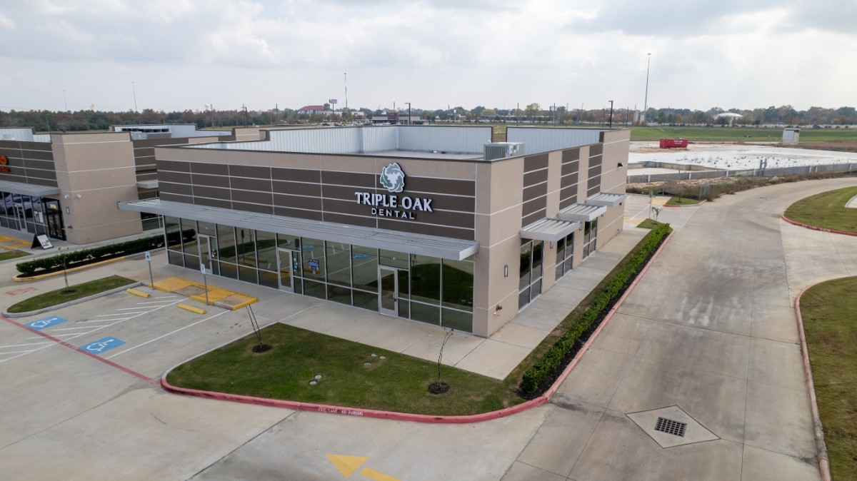 Modern one-story commercial building with 'Triple Oak Dental' sign, surrounded by empty parking spaces and a curving driveway under a cloudy sky.
