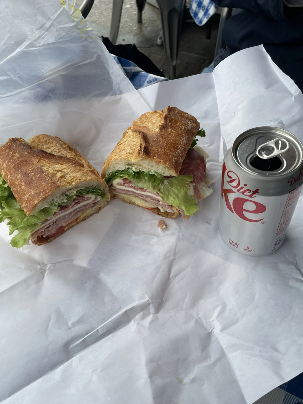 Sandwich with layers of deli meats and lettuce cut in half on white paper next to an open can of Diet Coke.