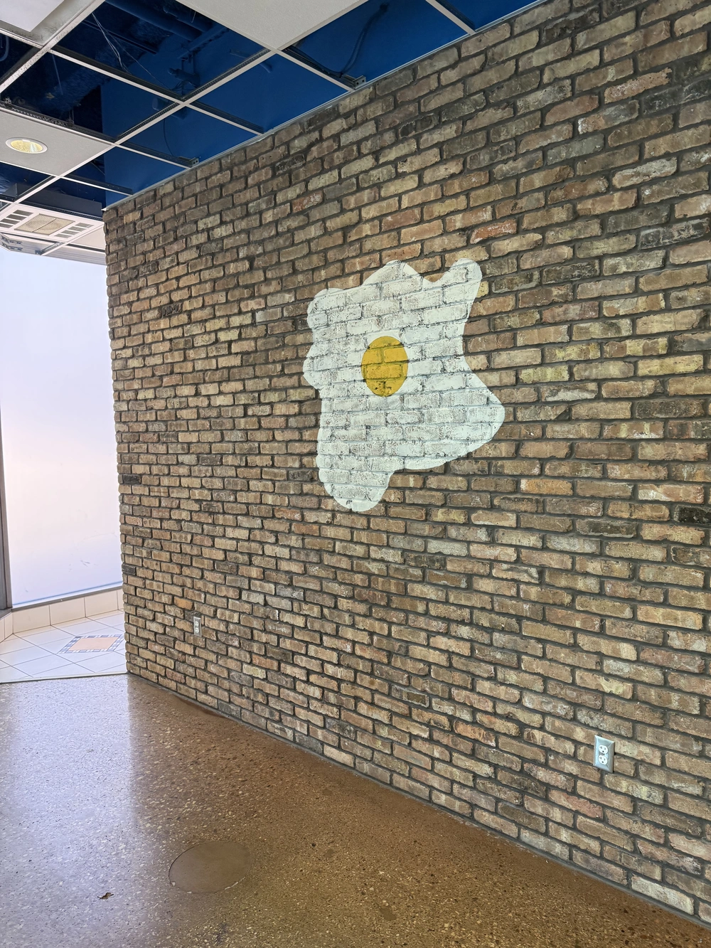 Brick wall with a painted white fried egg featuring a yellow yolk, next to a tiled floor area.