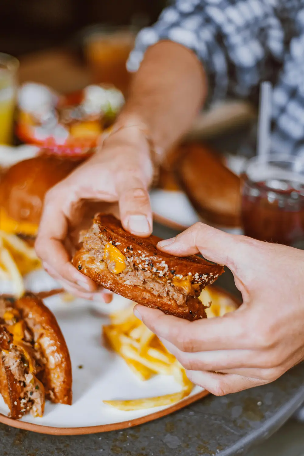 Two hands holding a cheeseburger with sesame seed bun and melted cheddar cheese, fries on plate in background.