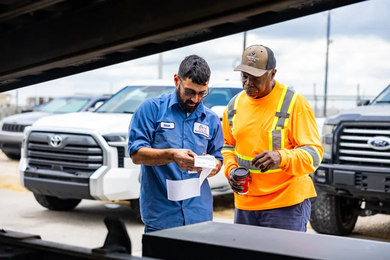 Mobile truck services technician Mike from Atlas Tire and Truck Center reviews paperwork with a worker beside fleet vehicles.