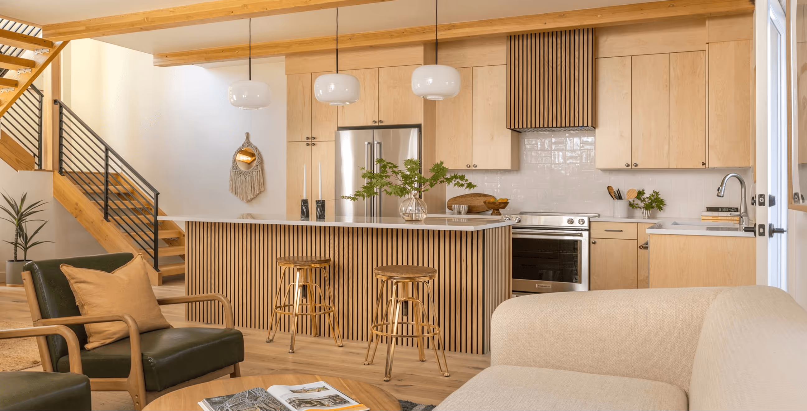 Modern kitchen with wood cabinets, kitchen island with two wooden stools, pendant lights, and seating area with green and beige chairs.