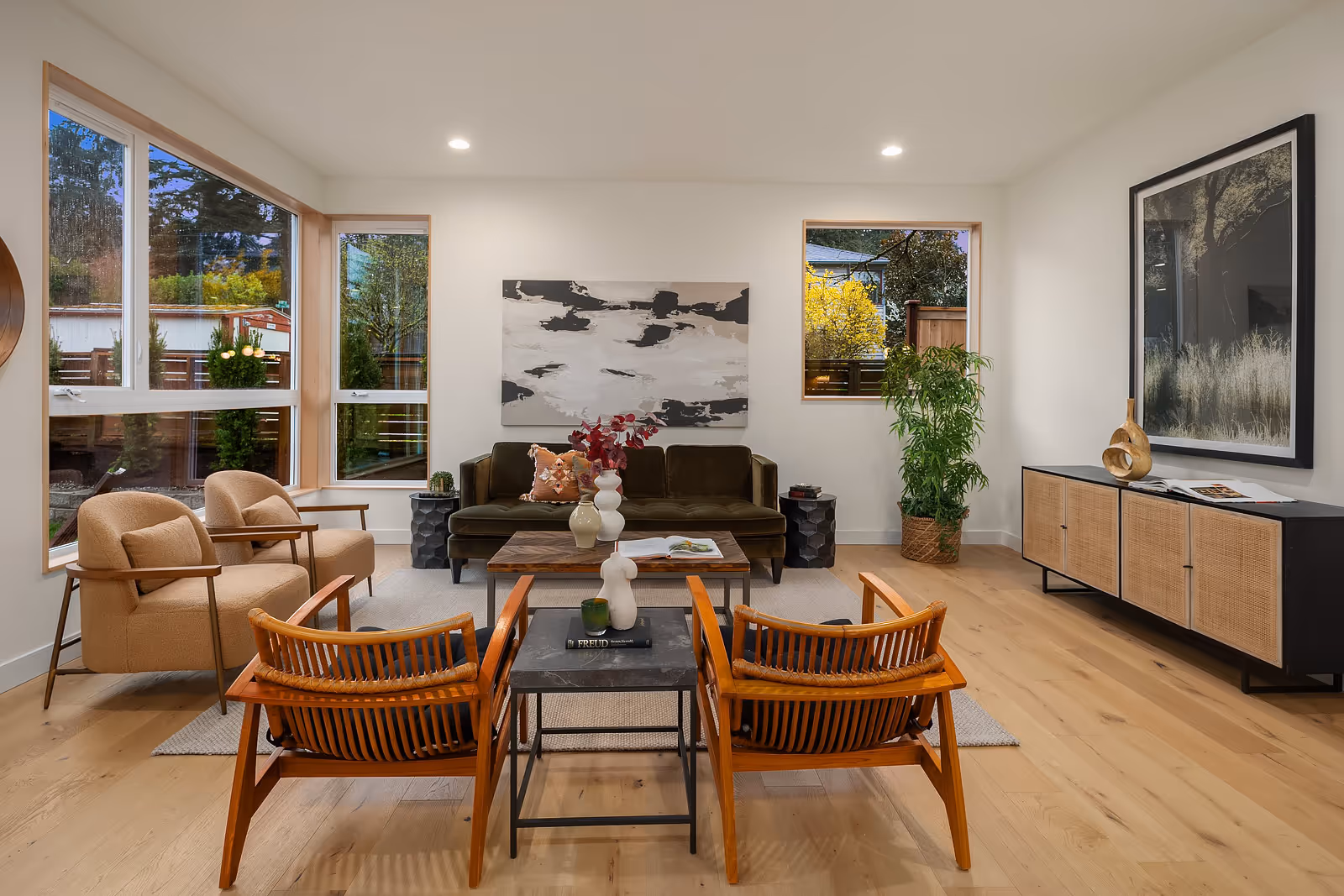 Modern living room with large windows, wooden floor, olive green sofa, two beige armchairs, two wooden chairs, black side tables, a plant, and abstract wall art.