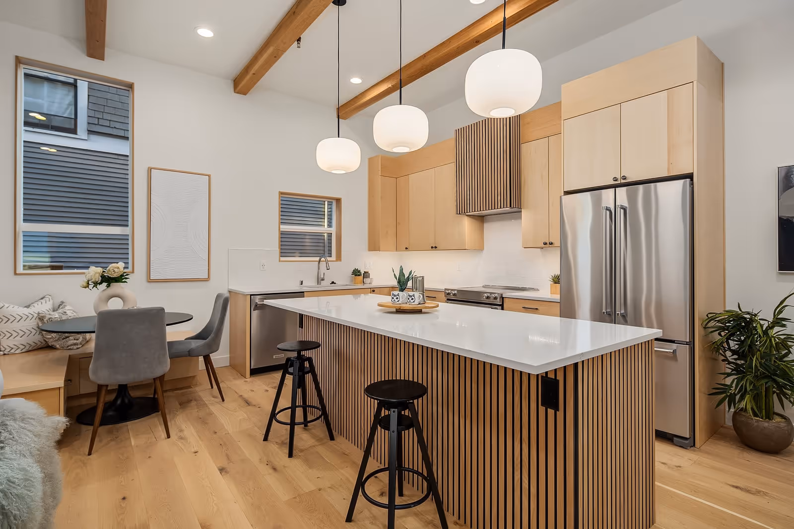 Modern kitchen with wooden cabinetry, white island countertop, three pendant lights, black stools, and a small dining nook with gray chairs.