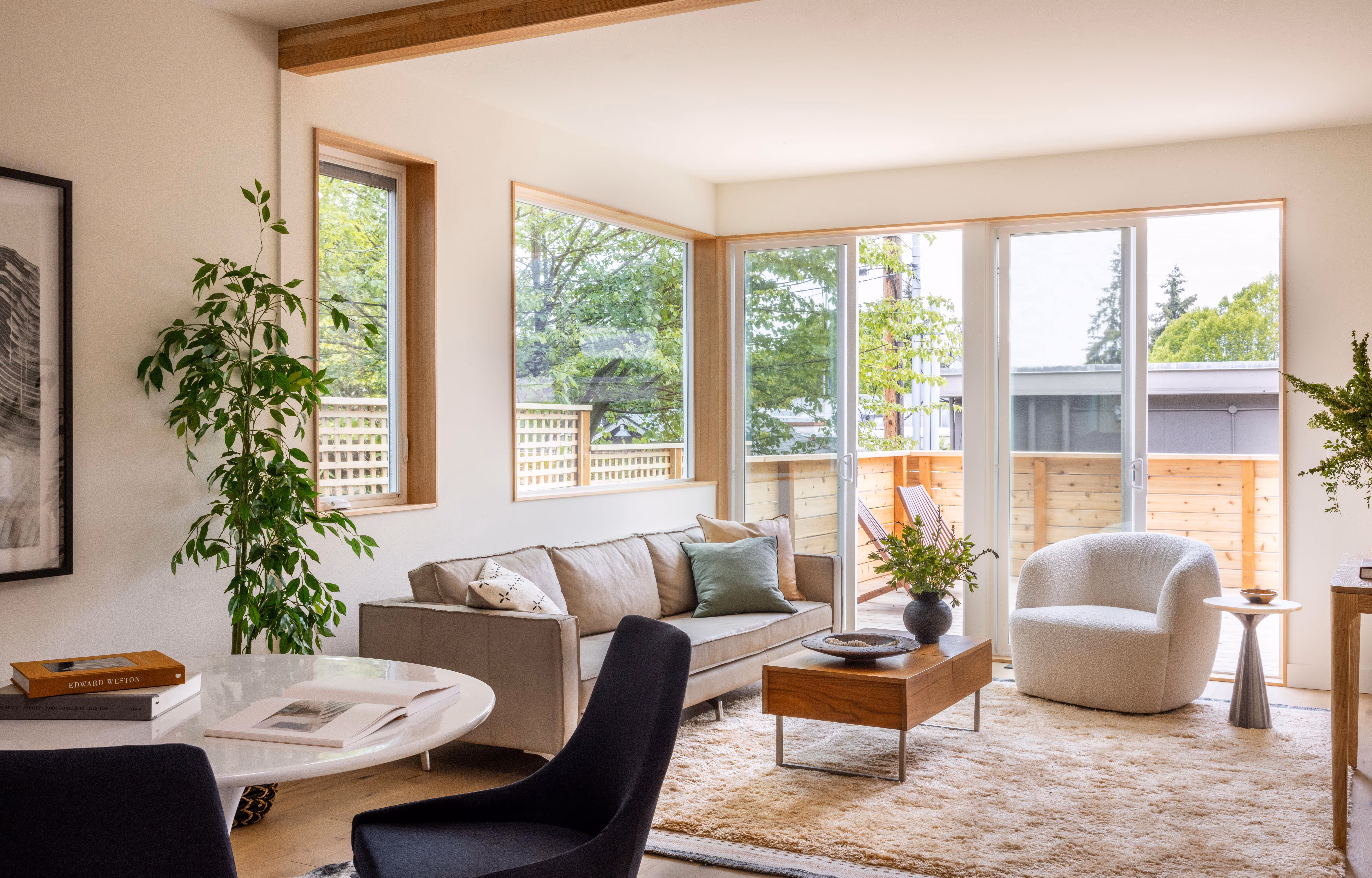 Bright living room with a beige sofa, white armchair, wooden coffee table, and large windows overlooking a wooden balcony and green trees.