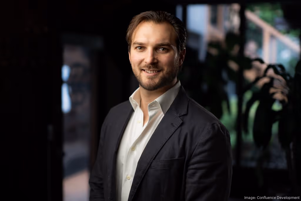 Smiling man in a white shirt and dark blazer standing indoors with blurred background.