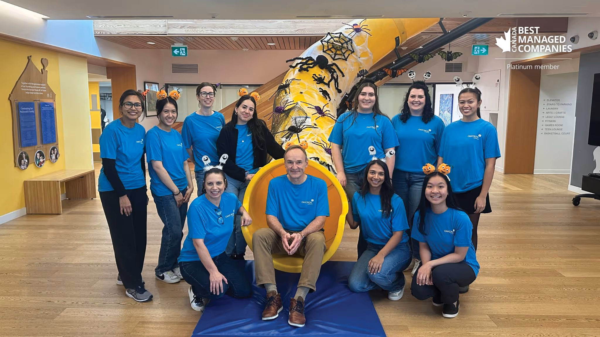 Group of eleven people wearing blue Omicron shirts posing indoors around a yellow slide decorated with Halloween spider and pumpkin decorations.