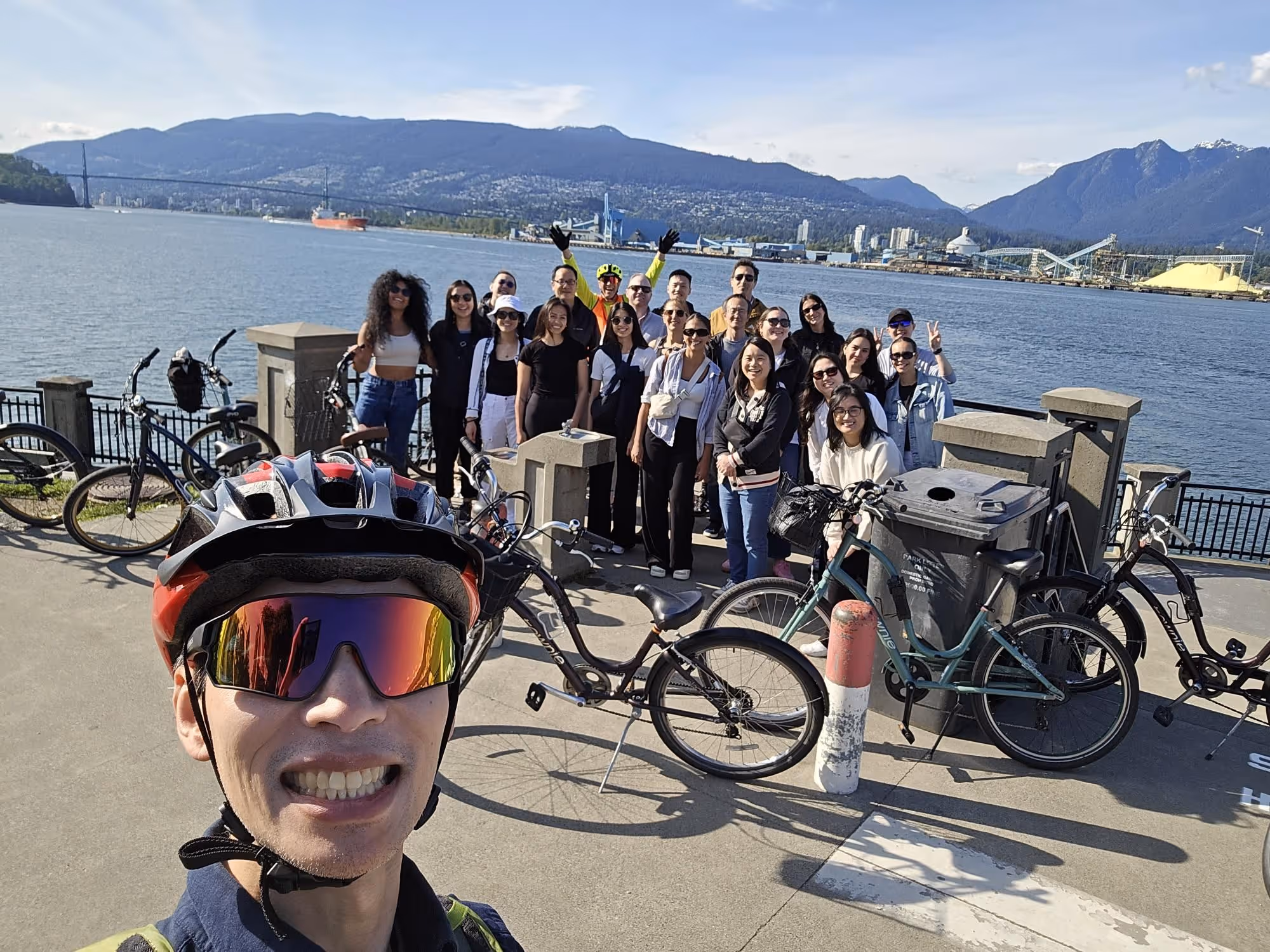 Group of people posing together near a waterfront with bicycles and mountains in the background on a sunny day.